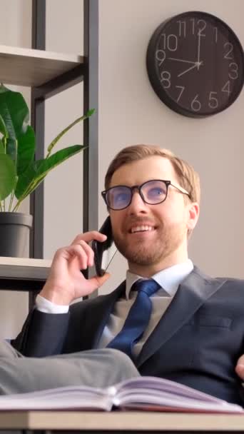 Un impiegato in giacca e cravatta che parla al cellulare e parla dei suoi successi, seduto in ufficio con i piedi sul tavolo. Video verticale - Filmati, video