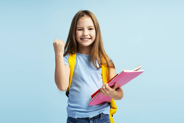 Smiling cheerful cute little girl wearing backpack holding books gesturing showing victory sign isolated on blue background. Back to school concept - Photo, Image