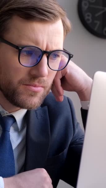 İş adamı takım elbisesiyle masada oturmuş dizüstü bilgisayara bakıyor ve ofisteki mali yatırım kararını düşünüyor. Dikey video - Video, Çekim