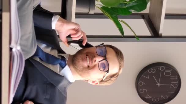 Trabajador de oficina en un traje hablando por teléfono móvil y hablando de sus logros, sentado en la oficina con los pies sobre la mesa. Vídeo vertical - Imágenes, Vídeo