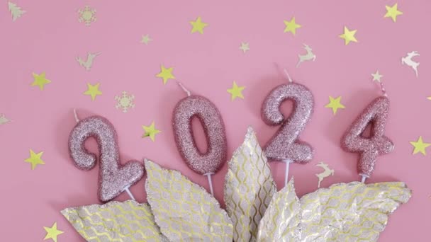 Az arany karácsonyi levelek, szétszórt konfetti és a 2024-es számú ünnepi csillogó gyertyák ága rózsaszín alapon fekszik, lapos fekvésű, körkörös fordulattal balra.. - Felvétel, videó