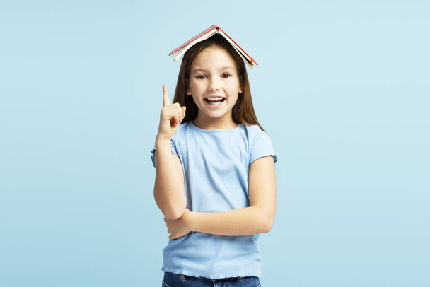 Sorrindo linda menina segurando livro na cabeça, apontando o dedo para cima, olhando para a câmera de pé isolado no fundo azul. Voltar ao conceito de escola - Foto, Imagem