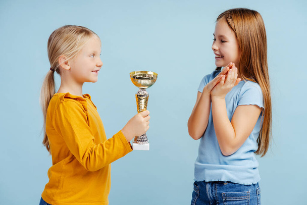 Roztomilá holčička dává vítězný pohár své sestře, stojící na modrém pozadí. Koncept vítězství, úspěšný - Fotografie, Obrázek