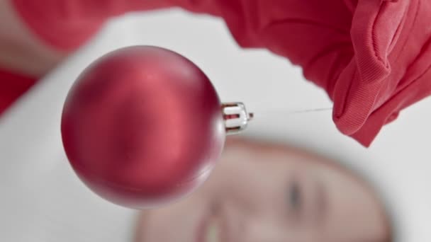 Pionowe wideo. Dekoracja choinki, czerwony balon w rękach dziewczyny trzymającej go w czerwonych rękawiczkach, wskazując kamerę przed nią i uśmiechając się. Koncepcja Bożego Narodzenia i Nowego Roku - Materiał filmowy, wideo