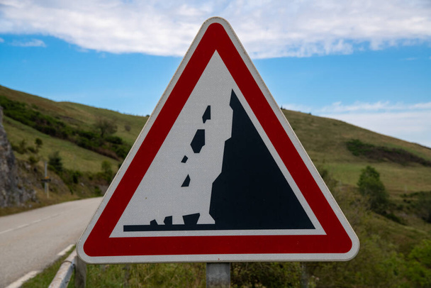 Μια τριγωνική πινακίδα μπροστά από ένα ορεινό φόντο στα χρώματα κόκκινο, λευκό και μαύρο, για να προειδοποιήσει για κατολισθήσεις, υποδεικνύεται από ένα βουνό και πέτρες που πέφτουν ως εικονόγραμμα - Φωτογραφία, εικόνα