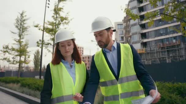 Contractors diversity engineers two people colleagues man woman builders architects wearing safety uniform protective helmets hardhats talking looking at blueprint building architecture plan in city - Footage, Video