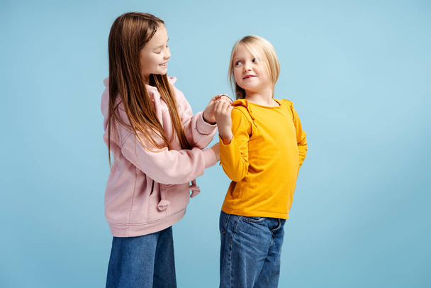 Portrait of smiling, attractive children, older sister brushing hair of younger, wearing stylish colored clothes standing isolated on blue background. Childhood concept, help, care - Photo, Image