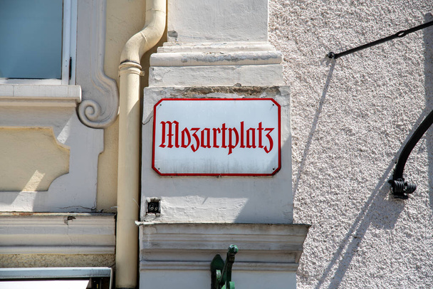 Egy fehér fémjel a Mozartplatz felirattal, régi német betűkkel. A tábla a ház falán lóg, piros betűkkel és piros szegéllyel.. - Fotó, kép