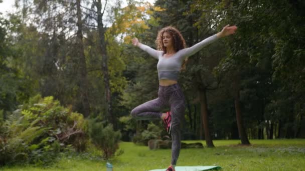 Кавказская тонкая мышечная гибкость спокойная спортивная женщина балансирует на одной ноге на коврике в парке летом фитнес девочка йог тренировки благополучия тренировки женщина стоит в позе дерева растягивая ногу перед - Кадры, видео