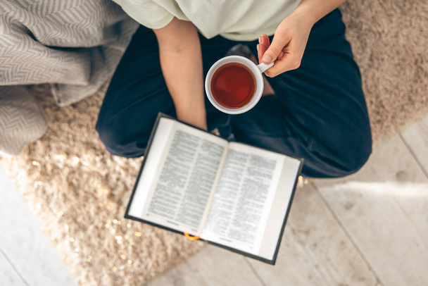 Vista dall'alto, donna che tiene una tazza di tè mentre legge un libro sul pavimento di un accogliente soggiorno. - Foto, immagini