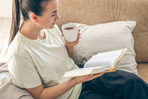 Young woman drinking hot tea, reading favorite bestseller novel poetry in paper book. Enjoying cozy lazy weekend holiday time, relaxing alone on sofa at home. - Photo, Image
