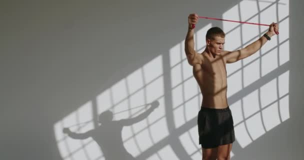 Atletický muž má cvičení a cvičení s odporovou páskou ve studiu s bílým pozadím. Samec bez trička s perfektními tělovými tréninkovými rameny a zádovými svaly pomocí sportovního vybavení. - Záběry, video