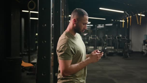 Američan Latino etnický muž procházení smartphone vážný atlet fit sport chlap osobní fitness trenér instruktor muž v tělocvičně kulturista sportovec pomocí mobilního telefonu při pohledu na kameru šťastný úsměv - Záběry, video