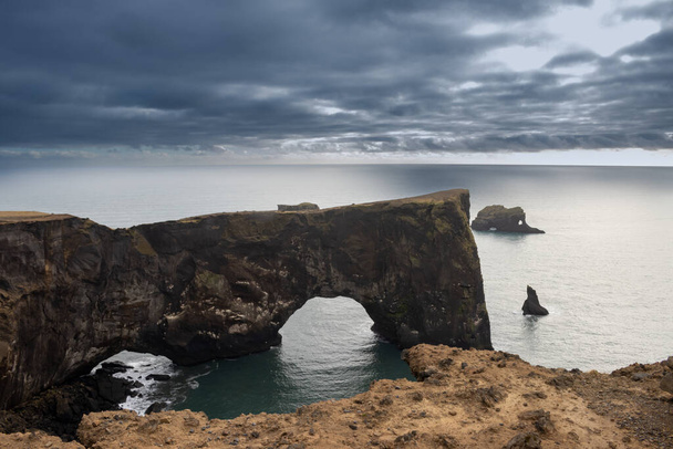 Διάσημη αψίδα Dyrholaey, παράθυρο σε ένα βράχο στον Ατλαντικό ωκεανό. Σύννεφο ουρανό το φθινόπωρο, αντανακλώντας κάποια ηλιοφάνεια μεταξύ των νεφών. Dyrholaey, Νότια Ισλανδία. - Φωτογραφία, εικόνα