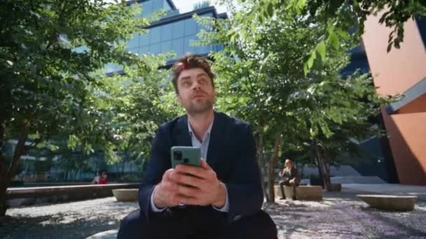Empleado viendo las redes sociales en el teléfono inteligente sentado soleado plaza verde de cerca. Un hombre de negocios relajado mandando mensajes de texto en el celular en el descanso laboral al aire libre. Hombre freelancer buscando mensaje de lectura de teléfono móvil. - Metraje, vídeo