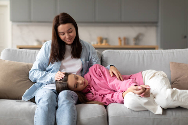 Tired daughter covered with warm blanket, lying on mother knees while relaxing together on couch at home, loving mom touching head of teen girl - Photo, Image