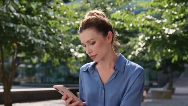 İş kadını akıllı telefon ekranında güneşli şehir parkında dinleniyor. Ciddi kadın serbest çalışan, yeşil ağaçlı bir ara sokakta oturan cep telefonu uygulamasına bakıyor. Kendine güvenen iş kadını mesajı okuyor.. - Video, Çekim