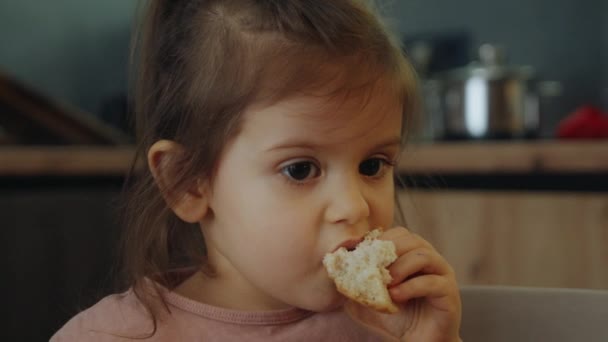 Feliz niña linda disfrutando de comer pan en el desayuno en la cocina en casa. Niña sentada en la mesa de la cocina temprano en la mañana y desayunando - Metraje, vídeo