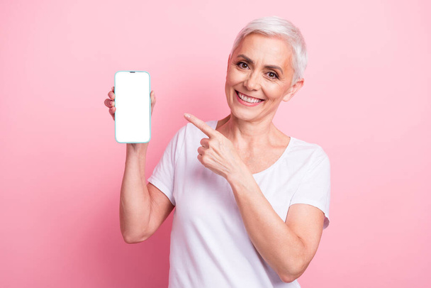 Photo of clever senior woman with short hair wear white t-shirt directing at smartphone screen empty space isolated on pink background. - Valokuva, kuva