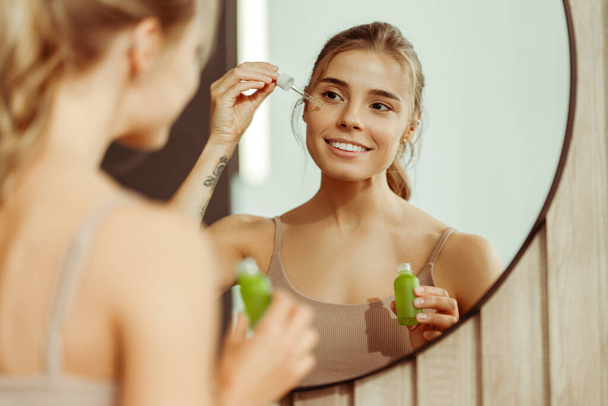 Piękna młoda kobieta trzymająca zieloną butelkę z serum na twarzy, patrząca w lustro, stojąca w łazience w domu. Pojęcie pielęgnacji skóry, zabiegu kosmetycznego, nawilżającego - Zdjęcie, obraz