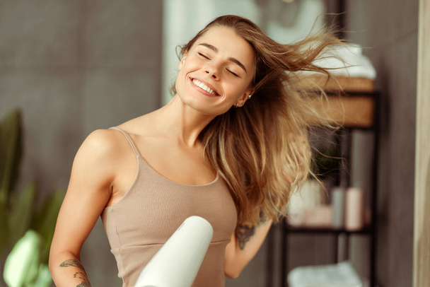 Feliz hermosa mujer joven secándose el pelo con secador, vistiendo camiseta de pie en el baño. Concepto de cuidado del cabello, peinado - Foto, Imagen