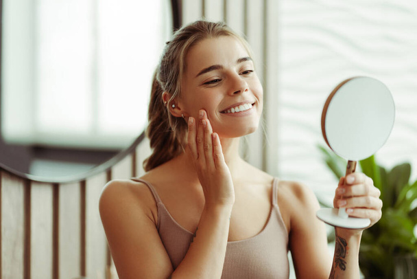 Atraente sorrindo mulher confiante segurando e olhando no espelho vestindo top tanque em pé no banheiro em casa. Conceito de cuidados com a pele, estilo de vida saudável - Foto, Imagem