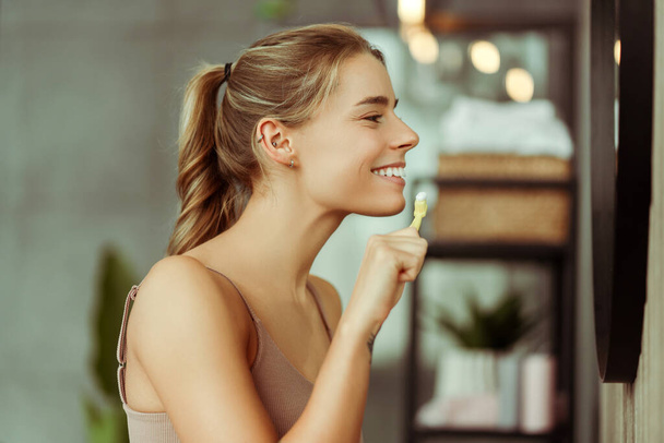 Uśmiechnięta młoda kobieta w koszulce ze szczoteczką do zębów z pastą, patrząca w lustro, myjąca zęby w łazience w domu. Pojęcie opieki stomatologicznej, leczenie - Zdjęcie, obraz