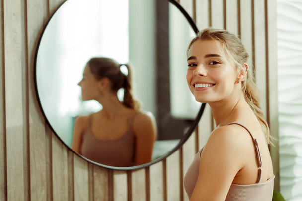 Πορτρέτο της θετικής όμορφη γυναίκα φορώντας μπλουζάκι, με τέλειο μακιγιάζ κοιτάζοντας κάμερα στέκεται στο μπάνιο στο σπίτι. Έννοια της φροντίδας του δέρματος, υγιεινό τρόπο ζωής - Φωτογραφία, εικόνα
