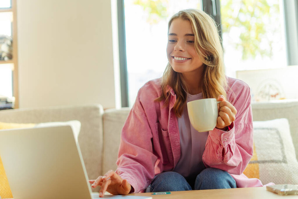 Portret młodej Amerykanki freelancerki trzymającej filiżankę kawy, siedzącej na kanapie, korzystającej z laptopa, piszącej na klawiaturze, sprawdzającej pocztę w kawiarni. ? oncept zdalnej pracy, technologii - Zdjęcie, obraz