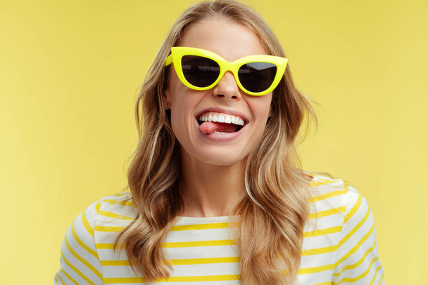 Portrét krásné usměvavé ženy se stylovými žlutými slunečními brýlemi ukazujícími jazyk ven, pohrávající si, stojící izolovaně na žlutém pozadí. Koncept reklamy - Fotografie, Obrázek