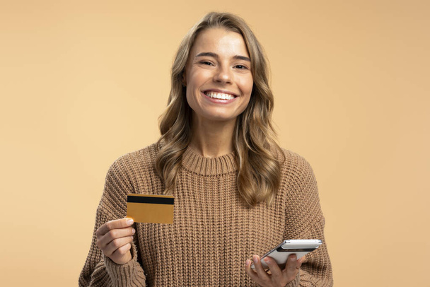 Porträt einer jungen Frau, die Handy und Kreditkarte in der Hand hält, mit einer mobilen App isoliert auf beigem Hintergrund. Konzept des Online-Shoppings - Foto, Bild
