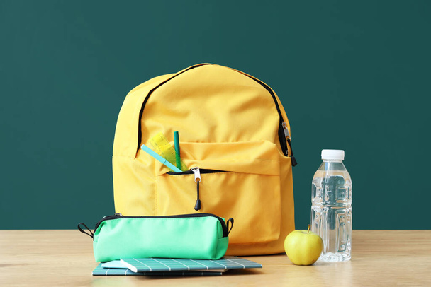 Gele schoolrugzak met briefpapier en flesje water op houten tafel bij groen schoolbord - Foto, afbeelding