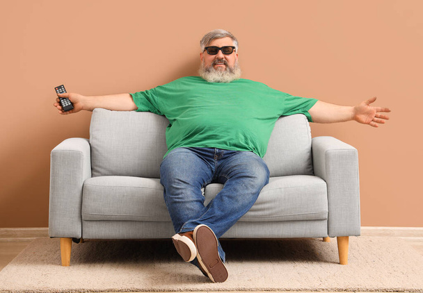 Uomo maturo con occhiali 3D che guarda la TV sul divano vicino alla parete beige - Foto, immagini