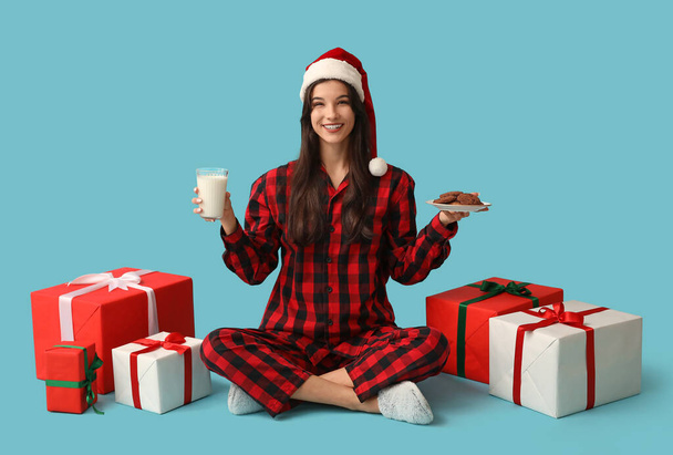 Beautiful young woman in checkered pajamas with sweet cookies, glass of milk and Christmas gift boxes on blue background - Photo, Image