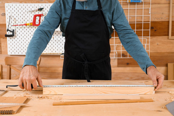 Mature carpenter measuring wooden plank at table in shop - Photo, Image