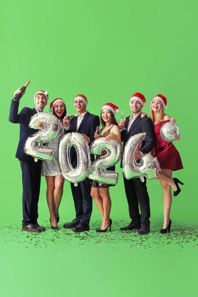 Grupa młodych ludzi w czapkach Mikołaja z rysunkiem 2024 i szampanem na zielonym tle. Święto Nowego Roku - Zdjęcie, obraz
