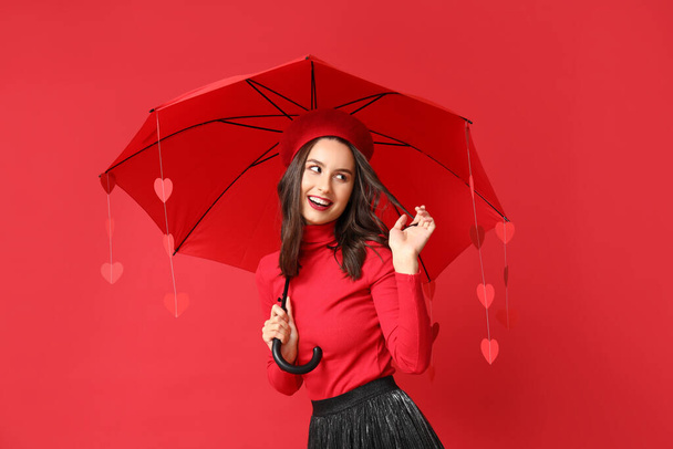 Beautiful young woman holding umbrella with paper hearts on red background. Valentine's Day celebration - Photo, Image
