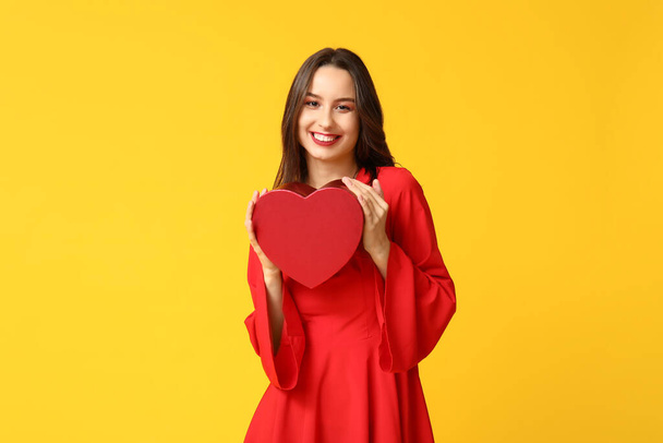 Красива молода жінка з подарунковою коробкою у формі серця на жовтому тлі. Святкування Дня святого Валентина - Фото, зображення