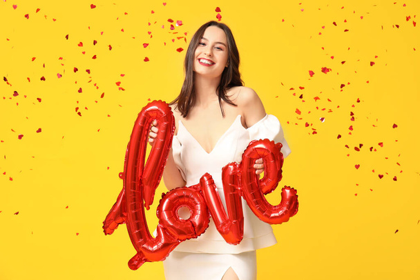 Beautiful young woman with air balloons in shape of word LOVE and confetti on yellow background. Valentine's Day celebration - Photo, Image