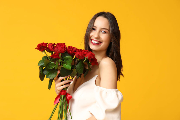 Beautiful young woman with bouquet of red roses on yellow background. Valentine's Day celebration - Photo, Image