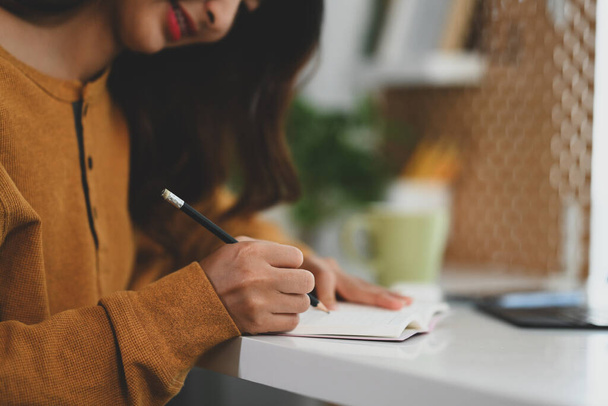 Tiro cortado de jovem mulher em suéter amarelo escrevendo em diário em sua mesa de trabalho. - Foto, Imagem