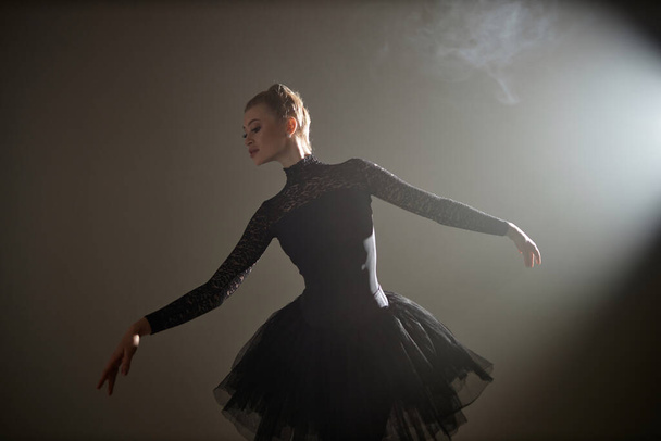 Young caucasian ballerina in black lace dress demonstrating ballet figure in dimly lit studio - Photo, Image