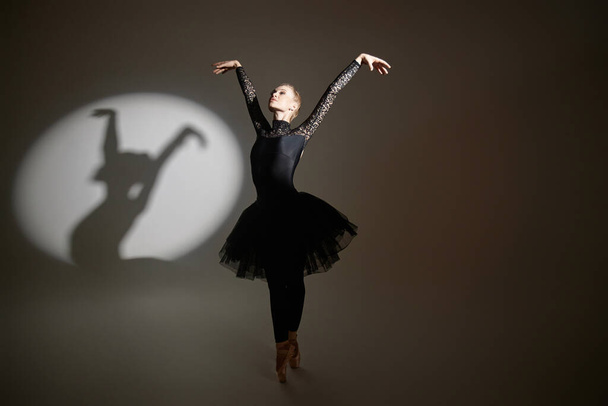 Ballerina che mostra la figura della danza durante le riprese fotografiche in studio, la sua ombra proiettata sullo sfondo a sinistra - Foto, immagini