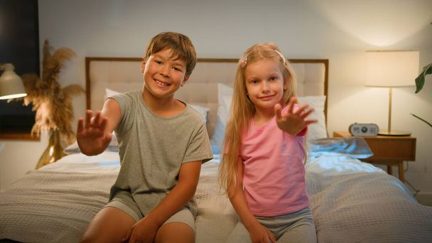 Dvě malé krásné přátelské kavkazské děti dítě děti chlapec dívka bratr sestra při pohledu kamera mává ahoj v ložnici na posteli vlny ruce pozdrav přítel nebo relativní komunikace prostřednictvím videohovoru doma - Fotografie, Obrázek