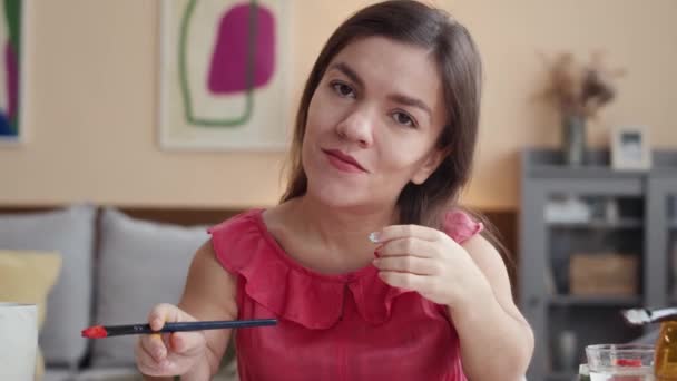 Portrait taille haute de jeune femme caucasienne tenant le pinceau à la main et regardant la caméra, assise au bureau dans le salon moderne avec des images abstraites lumineuses sur les murs et la peinture - Séquence, vidéo