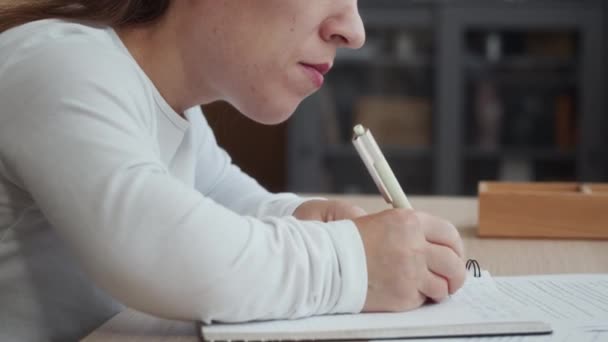 Tilt laukaus nuori valkoihoinen nainen lyhytkasvuisuus istuu pöydällä modernissa asunnossa ja kirjoittaa muistiinpanoja copybook tehdessään paperityötä - Materiaali, video