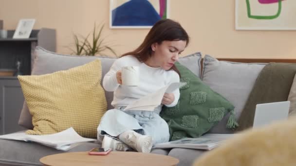 Νεαρή επιχειρηματίας με μικρό ανάστημα κάθεται στον καναπέ στο σύγχρονο σαλόνι, κοιτάζοντας μέσα από οικονομικές εκθέσεις και έγγραφα και τη χρήση φορητού υπολογιστή, ενώ εργάζονται από το σπίτι - Πλάνα, βίντεο