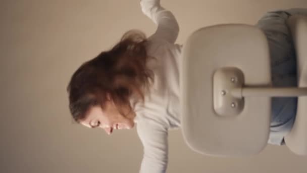 Vertical medium slowmo portrait of cheerful little woman spinning on office chair with her hands up and looking at camera - Footage, Video
