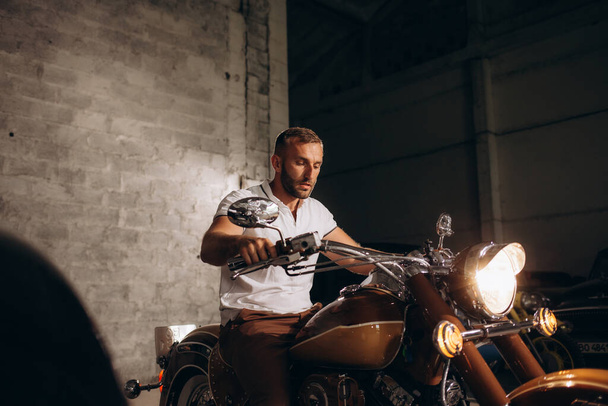 Un hombre se sienta en una motocicleta retro en su garaje - Foto, Imagen