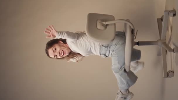 Vertical full length slowmo shot of young joyful Caucasian woman with dwarfism raising her hands and waving at camera while spinning on office chair on light studio background - Footage, Video